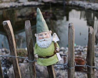 Puede incluir: Figura de gnomo de jardín con sombrero y túnica verdes, sosteniendo una pequeña pala. El gnomo tiene barba blanca y está colocado en una valla de madera con un estanque al fondo. La figura mide unos 15 cm de altura.