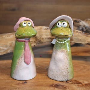 Könnte beinhalten: Zwei Keramik-Froschfiguren mit bemalten Details. Der Frosch links trägt einen rosa Hut und eine rosa Krawatte. Der Frosch rechts trägt einen rosa Hut und eine Perlenkette.