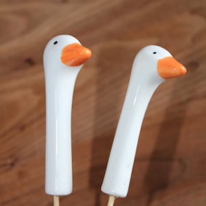 Könnte beinhalten: Zwei weiße Gänseköpfe aus Keramik, die an dünnen Holzstäben befestigt sind. Jeder Gänsekopf hat eine glatte weiße Oberfläche, einen leuchtend orangefarbenen Schnabel und einen kleinen schwarzen Punkt als Auge. Die Köpfe sind leicht zur Seite geneigt.