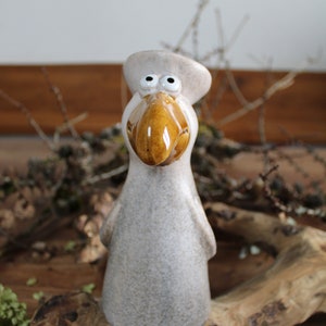 Könnte beinhalten: Keramik-Vogelfigur mit beigem Körper, braunem Schnabel und weißen Augen. Der Vogel trägt einen passenden beigen Hut. Die Figur steht auf einer Holzoberfläche mit trockenen Ästen im Hintergrund.