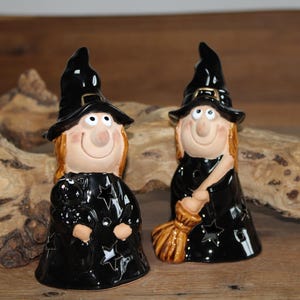 Könnte beinhalten: Zwei Keramik-Hexenfiguren. Jede Hexe hat einen schwarzen Hut, ein schwarzes Kleid mit Sternaussparungen und ein lächelndes Gesicht. Eine Hexe hält einen Besen. Die Figuren stehen auf einer Holzoberfläche, ideal für Halloween.