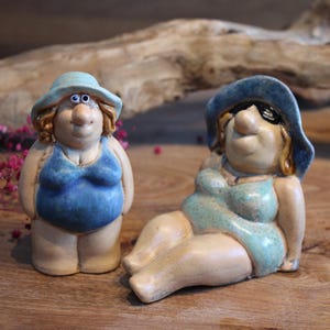 Könnte beinhalten: Zwei Keramikfiguren von Badegästen. Eine steht, die andere liegt. Beide tragen blaue Badeanzüge und Hüte. Die stehende Figur hat einen blauen Badeanzug und Hut. Die liegende Figur trägt eine Sonnenbrille sowie einen hellblauen Badeanzug und Hut. Die Figuren stehen auf einer Holzoberfläche.