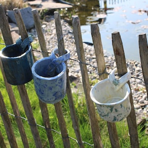 Könnte beinhalten: Drei Keramikbecher in Blau- und Weißtönen hängen an einem Holzzaun. Die Becher haben Henkel und sind mit Draht am Zaun befestigt. Der Hintergrund umfasst Gras und ein Gewässer.