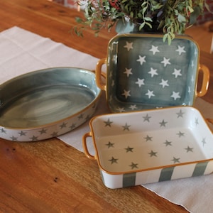 Könnte beinhalten: Drei Keramik-Backformen mit goldenen Griffen. Die Formen sind mit einem weißen Sternenmuster auf blauem Hintergrund verziert. Die kleinste Form hat einen weißen und blauen gestreiften Rand.