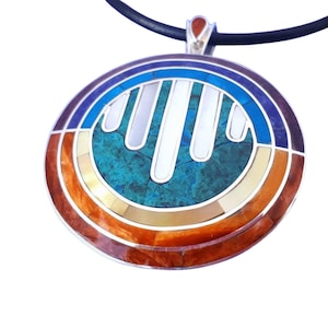 May include: A round pendant necklace with a silver-colored frame. The pendant features a turquoise center, surrounded by concentric rings of blue, white, orange, and brown. The necklace has a black cord and a small red accent at the top.