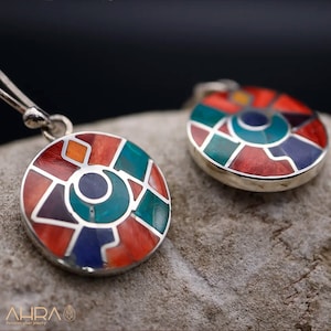 Puede incluir: Un par de pendientes de plata con un diseño circular. Los pendientes están hechos de plata y presentan un patrón geométrico colorido de piedras rojas, naranjas, amarillas, verdes, azules y moradas.