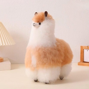 May include: A plush alpaca figurine with a soft, fluffy texture. The alpaca has a white body with patches of light brown fur on its back and head. It has small black eyes and ears, and a rectangular black nose.