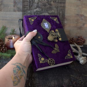 Può includere: Un diario rettangolare rilegato in tessuto di velluto viola intenso. La copertina presenta angolari in ottone decorati, un emblema ovale centrale raffigurante un simbolo della triplice luna e una chiusura in pelle nera con lucchetto in ottone.