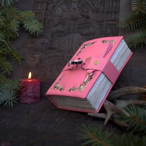 May include: A pink leather-bound journal with gold filigree corners and a clasp closure. The journal has a strap closure and a key charm. A lit red candle and evergreen branches are in the background, creating a mystical theme.