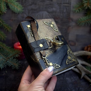 May include: A handcrafted journal with a black leather cover and gold accents. The journal features a gold-colored clasp and decorative corner pieces. The cover has a snakeskin pattern and a black leather strap. The journal is held by a person.