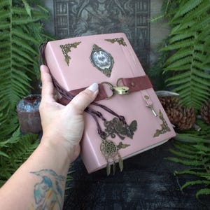 May include: A dusty pink journal featuring ornate brass corner accents, a central oval cameo, and a dark brown leather strap closure secured with a small brass padlock and key. Decorative charms including a moth and a dreamcatcher hang from the strap.