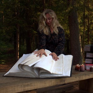 May include: A large, thick, open book with numerous blank, off-white pages resting on a wooden surface. The book features a dark cover and is positioned alongside three smaller, stacked books bound with black ribbons and three small, lit candles.