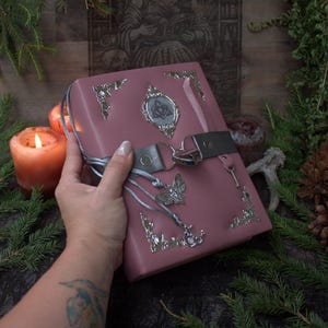 Puede incluir: Un diario de color rosa polvoriento con protectores de esquina de metal plateado, un emblema central plateado con un diseño de nudo en forma de corazón, un dije de mariposa plateado y un candado plateado en forma de corazón con una correa gris.