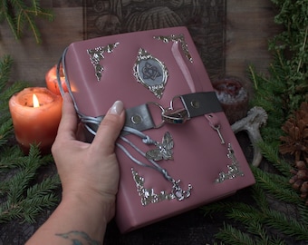 May include: A dusty rose-coloured journal featuring silver-toned metal corner protectors, a central silver emblem with a heart-shaped knot design, a silver butterfly charm, and a silver heart-shaped padlock secured by a grey strap across the cover.