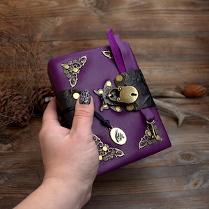May include: A purple leather journal with ornate brass corner accents and a decorative clasp. The journal features a black leather strap, a purple ribbon bookmark, and a small charm. The journal is closed.