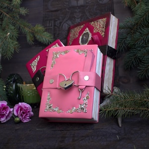 May include: A collection of decorative journals with ornate silver corner accents. The front journal is a vibrant pink with a heart-shaped lock and key closure. Other journals are red with gold filigree and black ribbon closures. The journals are on a dark wooden surface.