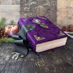 Pode incluir: Um livro revestido de veludo roxo com cantoneiras de latão ornamentadas e um emblema central. Uma tira de couro preta com fecho de latão prende o livro, e um pequeno pingente de pentagrama de latão pende de uma fita preta fina.