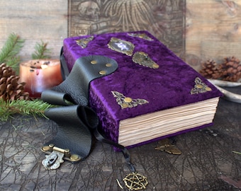 May include: A purple velvet-covered book featuring ornate brass corner protectors and a central decorative emblem. A black leather strap with a brass latch secures the book, and a small brass pentagram charm hangs from a thin black ribbon.