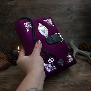 May include: A deep purple journal with silver embellishments and a black leather strap closure. The journal features a heart-shaped cameo, decorative corners, and a key charm. The cover is made of a soft, velvety material.
