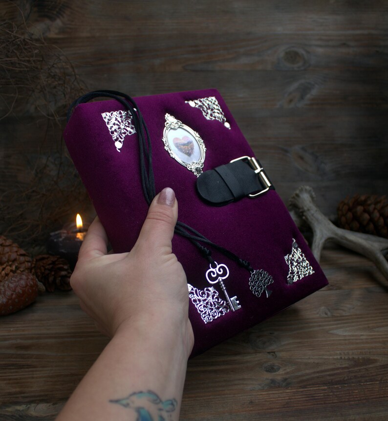May include: A deep purple journal with silver embellishments and a black leather strap closure. The journal features a heart-shaped cameo, decorative corners, and a key charm. The cover is made of a soft, velvety material.