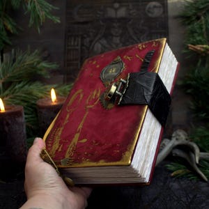 May include: A handmade, red-covered book with gold accents and a black leather strap. The cover features gold-colored utensils and decorative hardware. The book is held in a hand, with lit candles and greenery in the background.