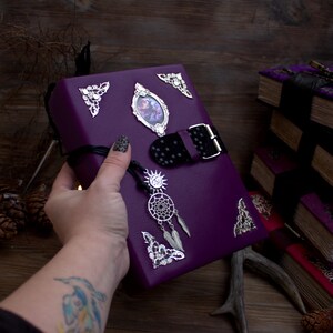 May include: A purple book with silver embellishments, including corner accents, a framed image, and a buckle closure. A silver dreamcatcher charm hangs from the book. The book is held in a hand with a tattoo and nail art.