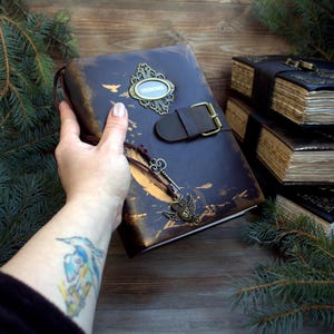 May include: A dark blue leather journal with a decorative metal clasp and a brown leather strap. The journal has a key and owl charm attached. Other journals are stacked in the background. The journal is held by a person.