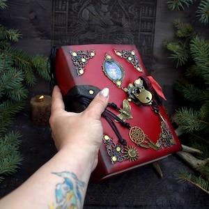 May include: A red leather-bound journal with ornate metal corner accents and decorative charms. The journal features a butterfly, dreamcatcher, and sun pendant. A black cord wraps around the spine, and a small lock secures the journal.