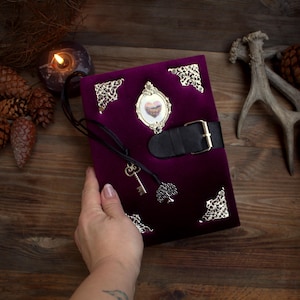 May include: A deep purple velvet journal with ornate silver corner accents and a decorative frame. The journal features a black leather strap with a buckle closure and a key charm. A small tree charm hangs from the journal.