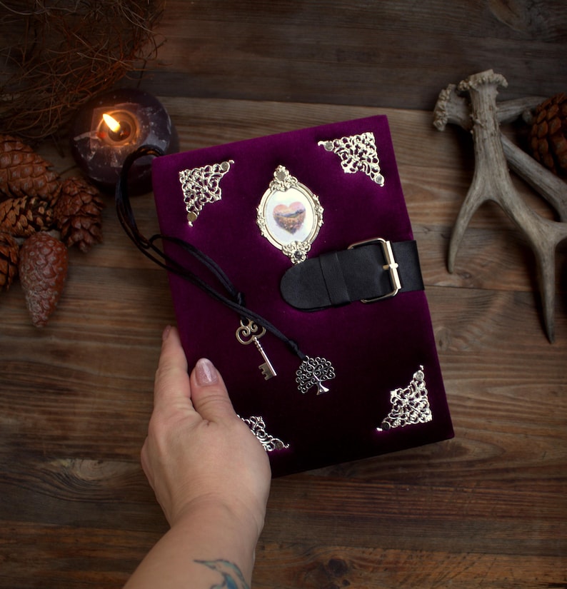 May include: A deep purple velvet journal with ornate silver corner accents and a decorative frame. The journal features a black leather strap with a buckle closure and a key charm. A small tree charm hangs from the journal.