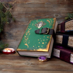 May include: A green leather-bound journal with gold accents and a decorative clasp. The cover features gold star and leaf designs. A brown leather strap with a buckle secures the journal. Other journals are stacked in the background.