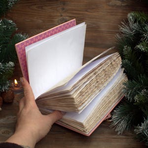 May include: An open, pink-covered journal with aged, cream-colored pages. The journal's spine is visible, revealing a patterned pink and white design. A hand holds the pages open, with a lit candle and pine cones in the background, suggesting a cozy, vintage aesthetic.