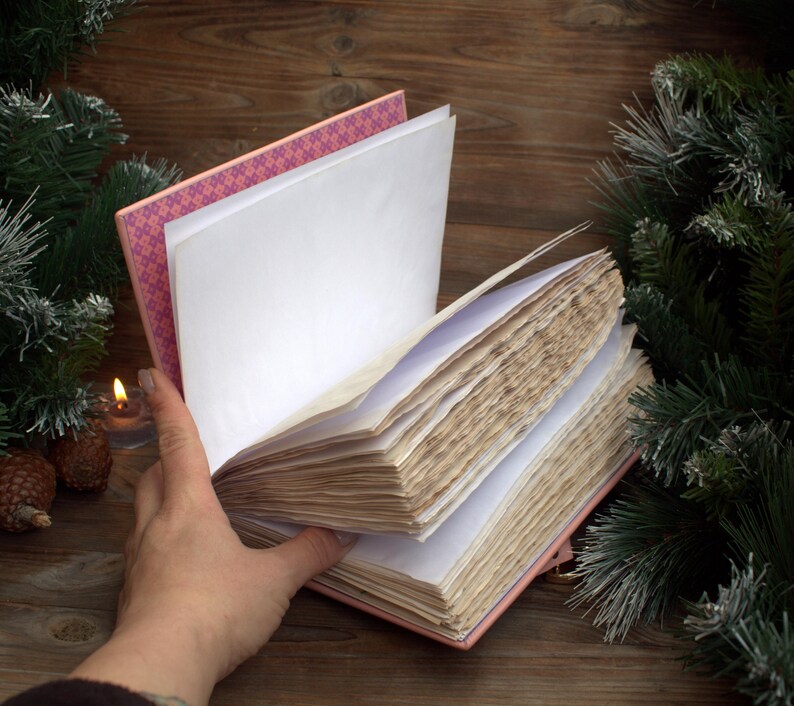 May include: An open, pink-covered journal with aged, cream-colored pages. The journal's spine is visible, revealing a patterned pink and white design. A hand holds the pages open, with a lit candle and pine cones in the background, suggesting a cozy, vintage aesthetic.