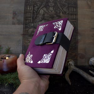 May include: A deep purple velvet journal featuring four ornate silver-toned metal corner protectors and a black leather strap closure with a silver-toned metal buckle. The journal contains numerous white paper pages bound within the velvet cover.