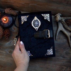 May include: Black velvet journal with silver corner accents and a decorative oval frame featuring a moon phase design. A black leather strap with a gold buckle secures the journal. A gold key hangs from a black cord.