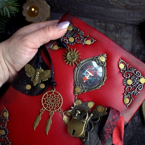 May include: Red leather journal with ornate metal embellishments, including a sun, a butterfly, and a dreamcatcher. The journal has a lock and key closure and decorative corner pieces. The cover is a vibrant red.
