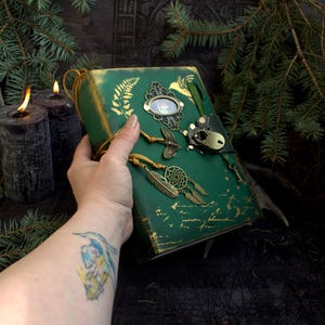 May include: A handcrafted green journal with gold accents, featuring a fern design and decorative metal charms. The journal has a leather strap closure and a dreamcatcher charm. The cover is embellished with a vintage-style frame and dragonfly charm.
