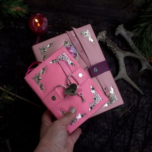 May include: Pink leather journals with silver corner accents and heart-shaped lock closures. One journal is held in a hand, while another is secured with a purple strap. A lit candle and deer antlers are in the background.