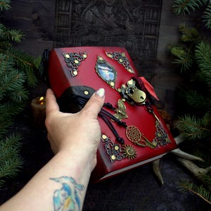 May include: A red leather-bound journal with ornate brass corner accents and a decorative lock. The cover features a butterfly charm, a dreamcatcher, and a sun pendant. The journal is held in a hand.