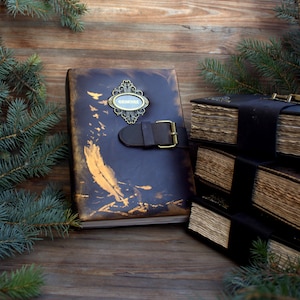 May include: A dark brown leather-bound journal with a decorative metal clasp and the word "GRIMOIRE" on a small plaque. The cover has gold accents and a leather strap with a buckle. Additional journals are stacked in the background.