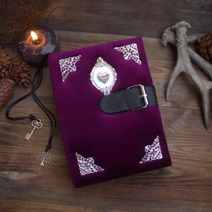 May include: A deep purple velvet journal with silver filigree corner details and an oval frame. It has a black leather strap with a silver buckle. The oval frame contains a small heart-shaped image.