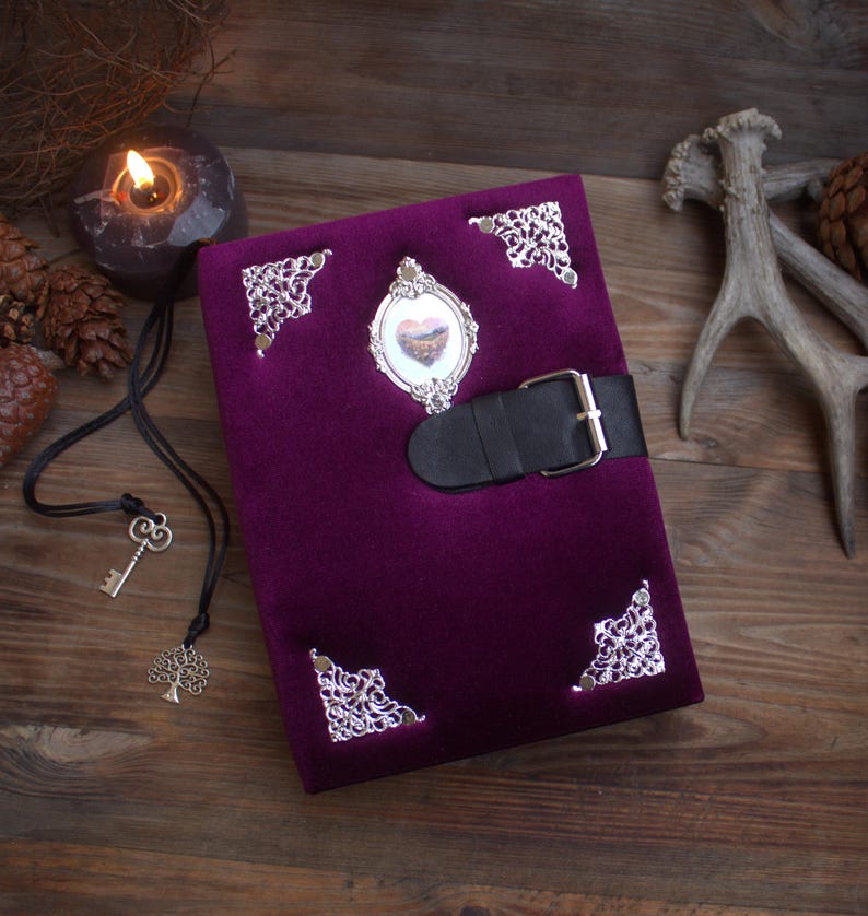 May include: A deep purple velvet journal with silver filigree corner details and an oval frame. It has a black leather strap with a silver buckle. The oval frame contains a small heart-shaped image.