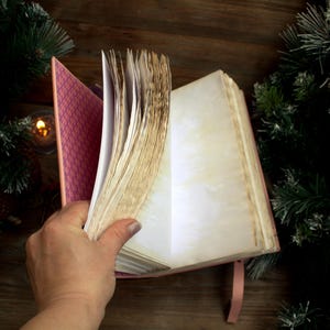 May include: A pink journal with aged, cream-colored pages is held open, revealing blank pages. The journal's cover has a patterned interior. A pink ribbon bookmark is visible. The background includes evergreen branches and a candle.