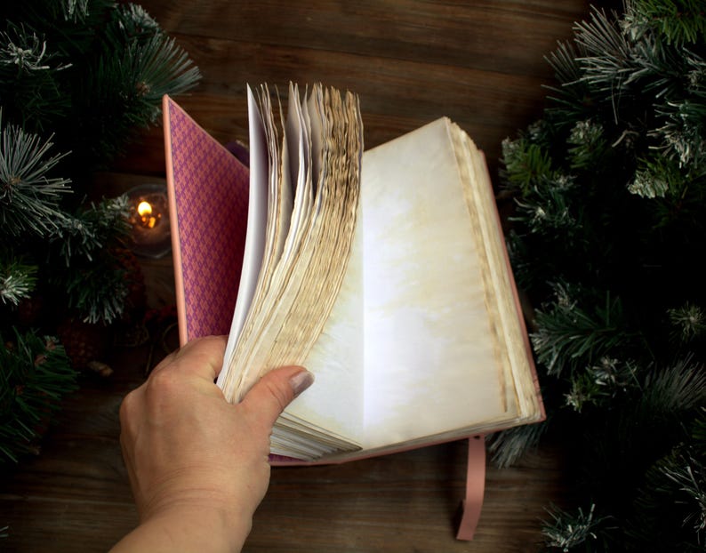 May include: A pink journal with aged, cream-colored pages is held open, revealing blank pages. The journal's cover has a patterned interior. A pink ribbon bookmark is visible. The background includes evergreen branches and a candle.