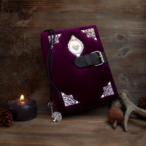 May include: A deep purple velvet journal with silver corner detailing and a black leather clasp. A silver key pendant hangs from the journal. A lit candle, pine cones, and a deer antler are in the background.