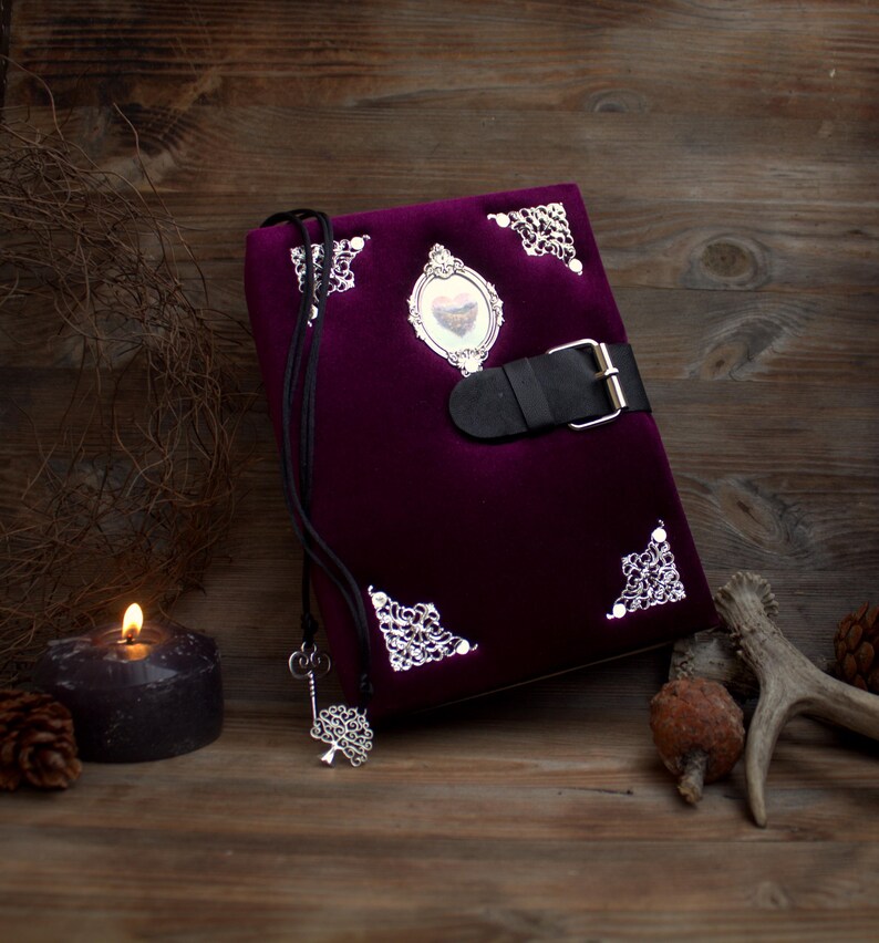May include: A deep purple velvet journal with silver corner detailing and a black leather clasp. A silver key pendant hangs from the journal. A lit candle, pine cones, and a deer antler are in the background.