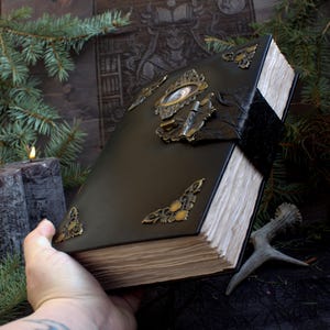 May include: A dark green leather-bound book with ornate brass corner pieces and clasps. The book has aged, off-white pages and a black velvet closure. A lit candle and evergreen branches are in the background.