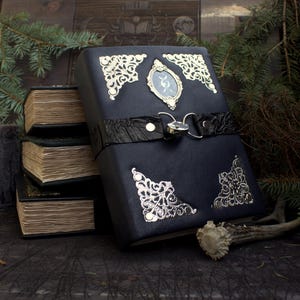 May include: A dark blue leather-bound journal with ornate silver corner accents and a central oval emblem. The journal features a black leather strap with a silver clasp. Several smaller, aged books are stacked nearby, suggesting a theme of mysticism or historical study.