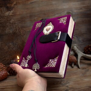 May include: A purple velvet journal with a black leather strap and buckle closure. The journal has decorative metal corner accents, a small framed image, and two pendant charms. The journal is held in a person's hand.