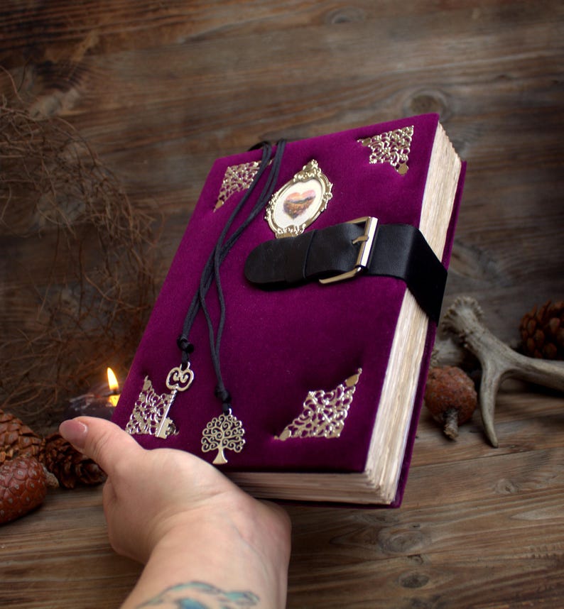 May include: A purple velvet journal with a black leather strap and buckle closure. The journal has decorative metal corner accents, a small framed image, and two pendant charms. The journal is held in a person's hand.